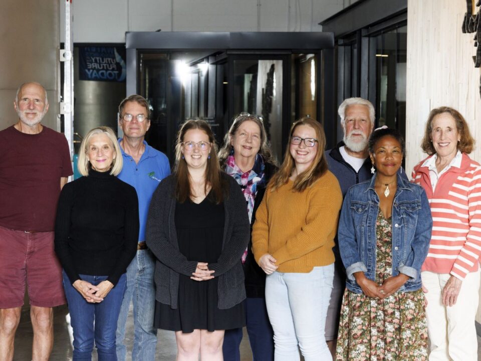 Group of nine people smiling and posing for a photo in a modern building. They are diverse in age and are dressed in casual and semi-casual attire. 