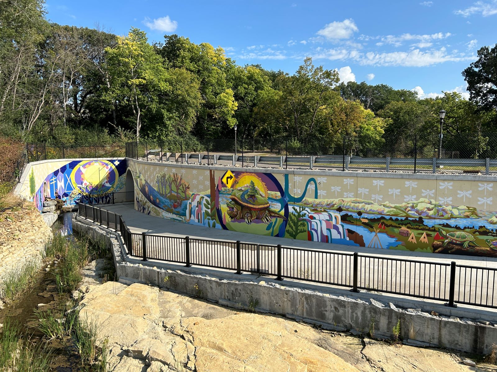 Colorful mural with vibrant aquatic and wildlife motifs painted on a curved wall along a paved pathway next to a stream channel, below a treelined road.