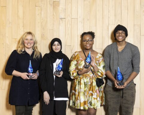 Four smiling people stand posing for a photo while they are each holding a blue-swirled glass raindrop award.