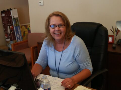 A smiling woman looks up at the camera while sitting in front of a computer. 