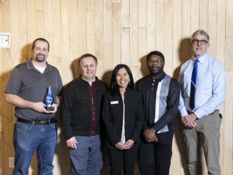 Five smiling people pose for a photo, the person on the left is holding a blue-swirled glass raindrop award.